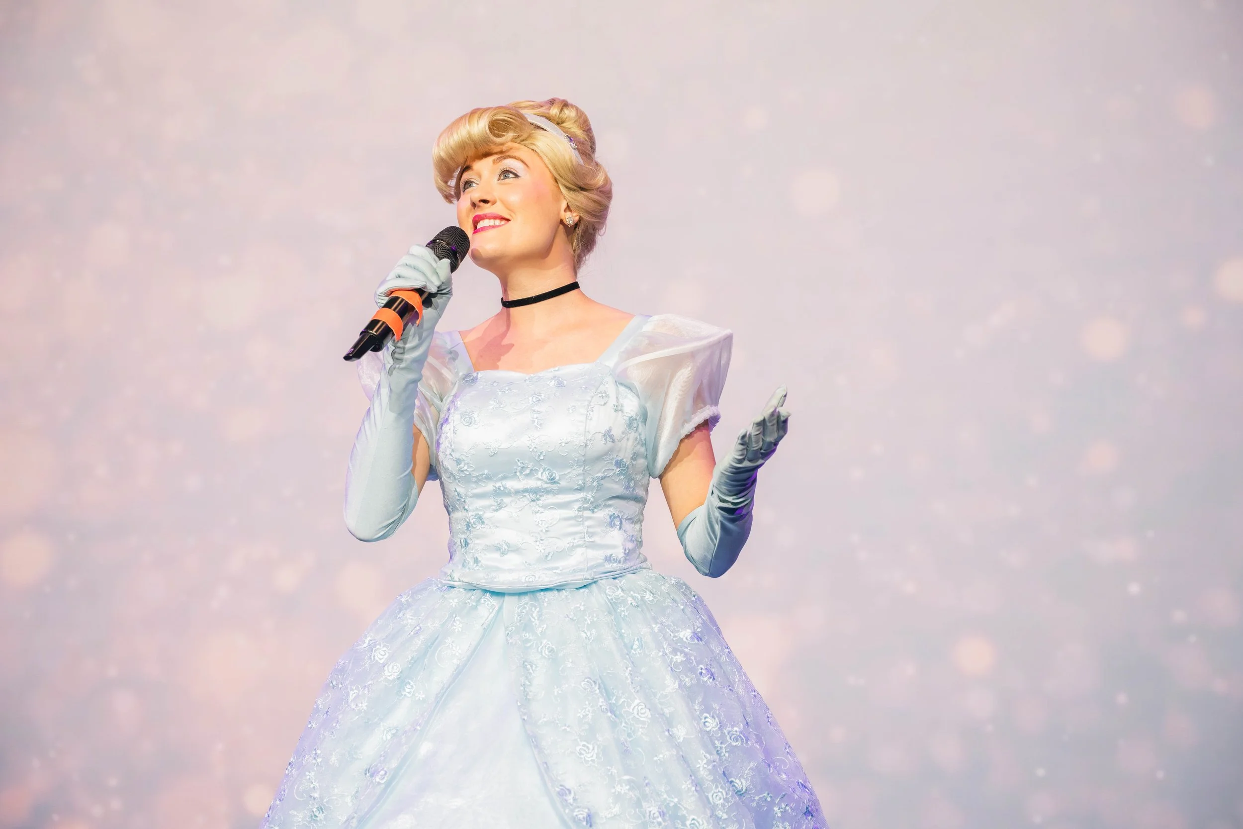 A woman dressed as Cinderella holding a microphone and smiling, with a dreamy sky background.