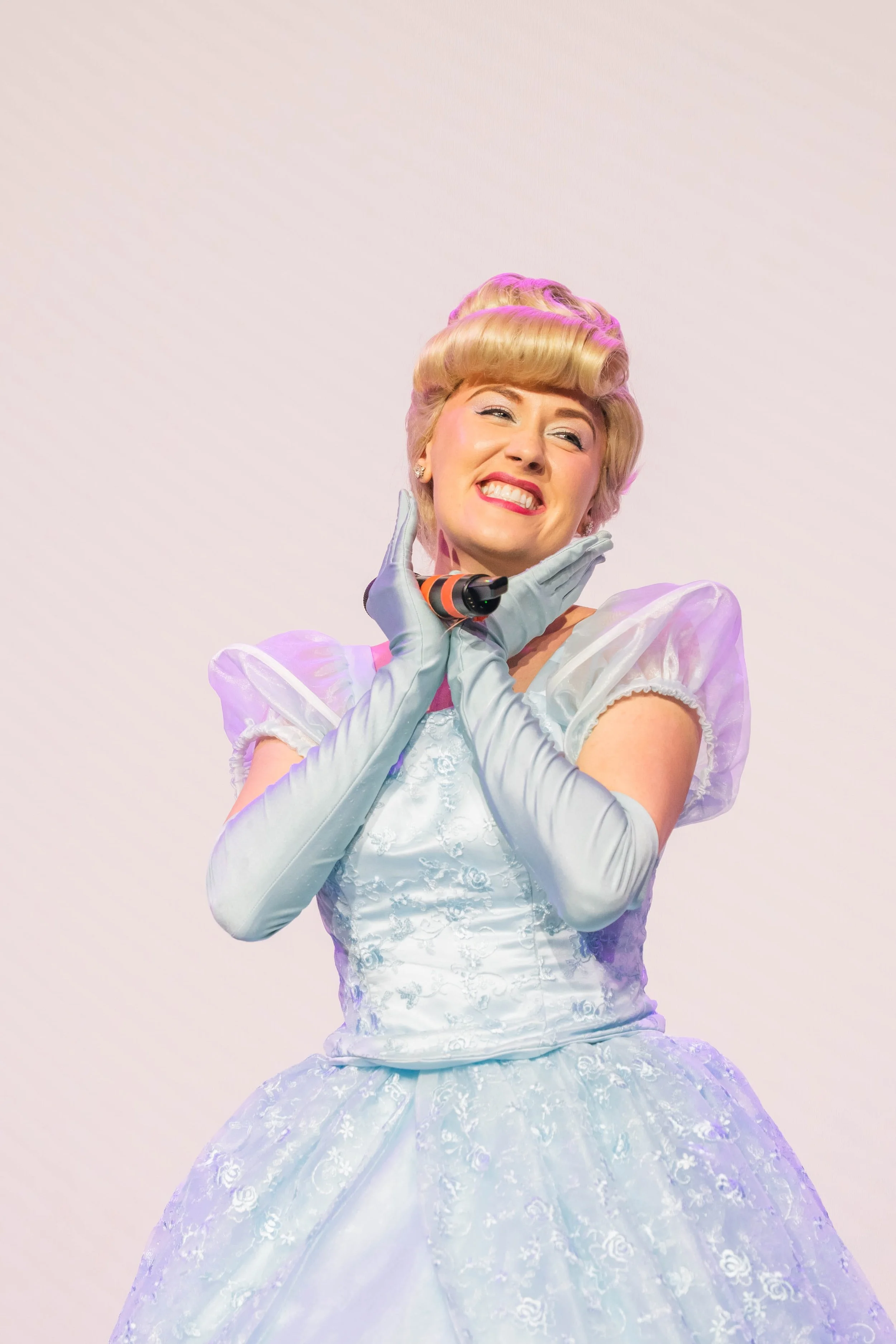 Woman dressed as Cinderella with a vintage hairstyle, wearing a light blue gown with puffed sleeves and white gloves, smiling and holding a black and orange hairbrush