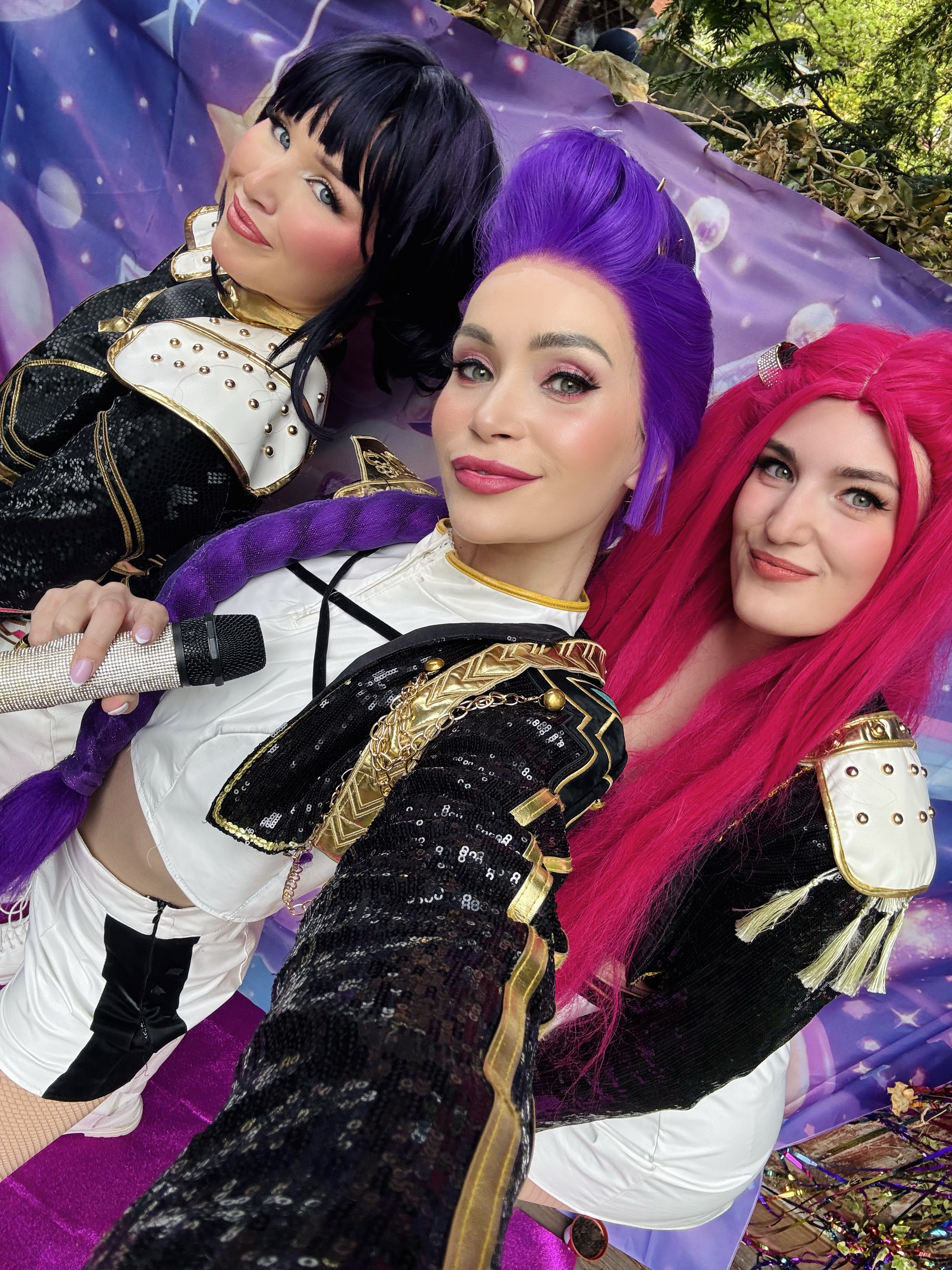 Three women dressed in colorful costumes with metallic and sequined details take a selfie together. They have vibrant hair colors: black, purple, and pink, and they are smiling at the camera. Behind them, there is a purple backdrop with star patterns