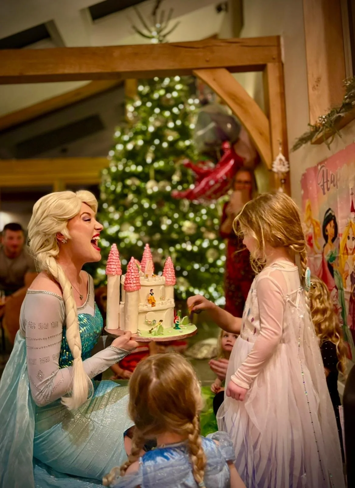 A woman dressed as Elsa from Frozen holding a castle-themed birthday cake with pink turrets, at a child's birthday party with a decorated Christmas tree in the background.