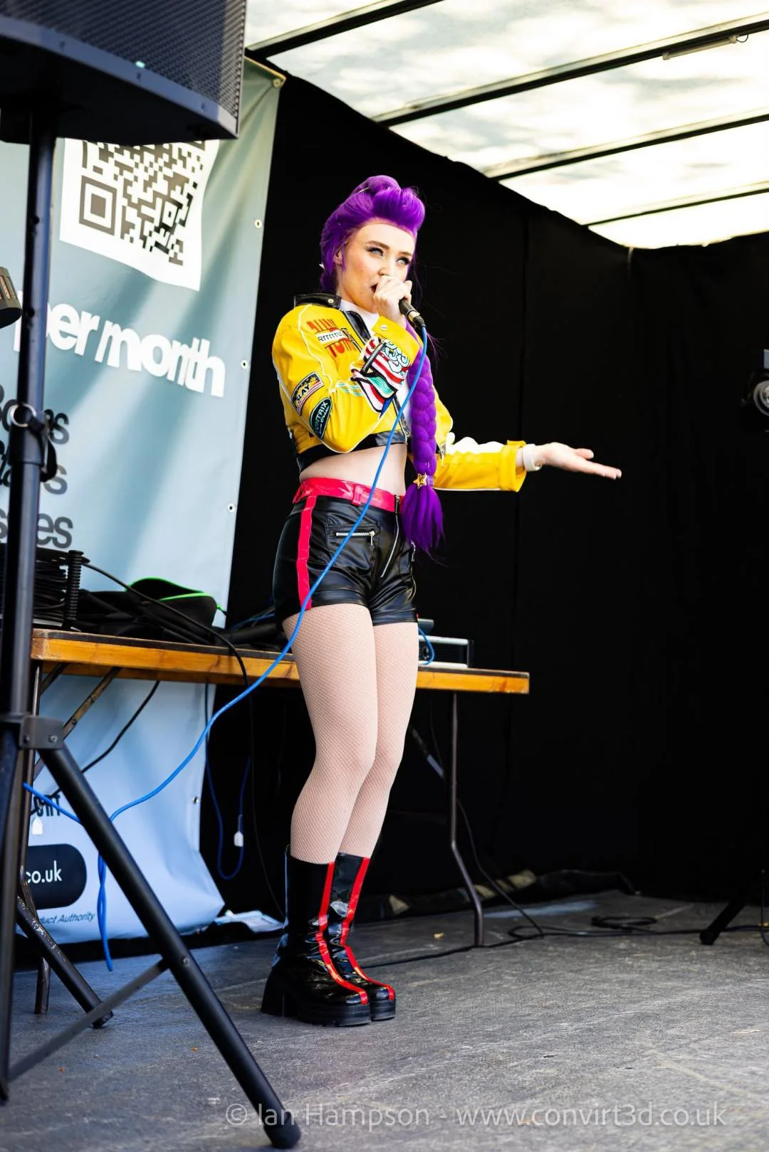 A woman performing on stage, with purple hair styled in a twisted braid, wearing a colorful jacket, black shorts, fishnet stockings, and platform boots, holding a microphone.