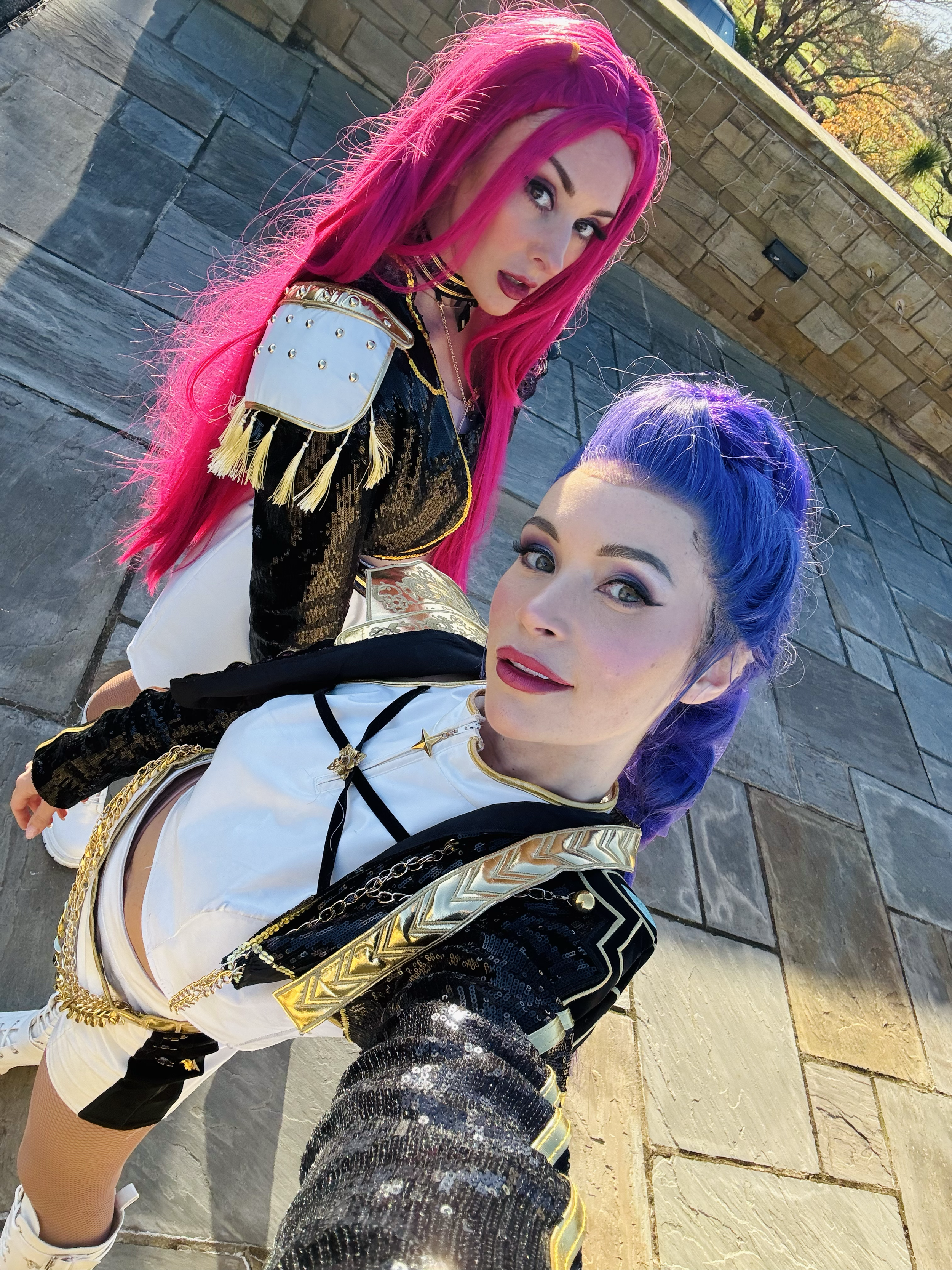 Two women in shiny, glamorous outfits with colorful hair, posing outdoors on a stone pavement with trees and a wall in the background.