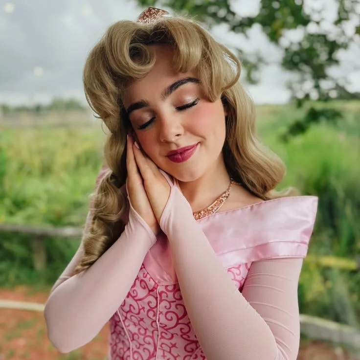 A woman dressed as Princess Aurora from Sleeping Beauty, wearing a pink gown, with long blonde hair styled in vintage curls, resting her face on her hands with eyes closed outdoors in a lush green setting.