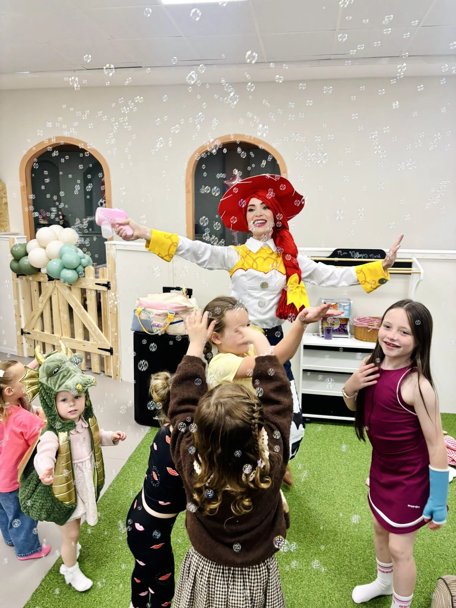 Children at a birthday party with a woman dressed as a character, surrounded by soap bubbles, some children are wearing costumes, and there are decorations including balloons and a small blackboard, on a green carpeted area.