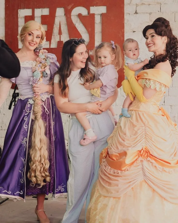 Four women, including Disney princesses Rapunzel and Snow White, hold young children at an event with a brick wall background featuring the word 'EAST' painted on it.