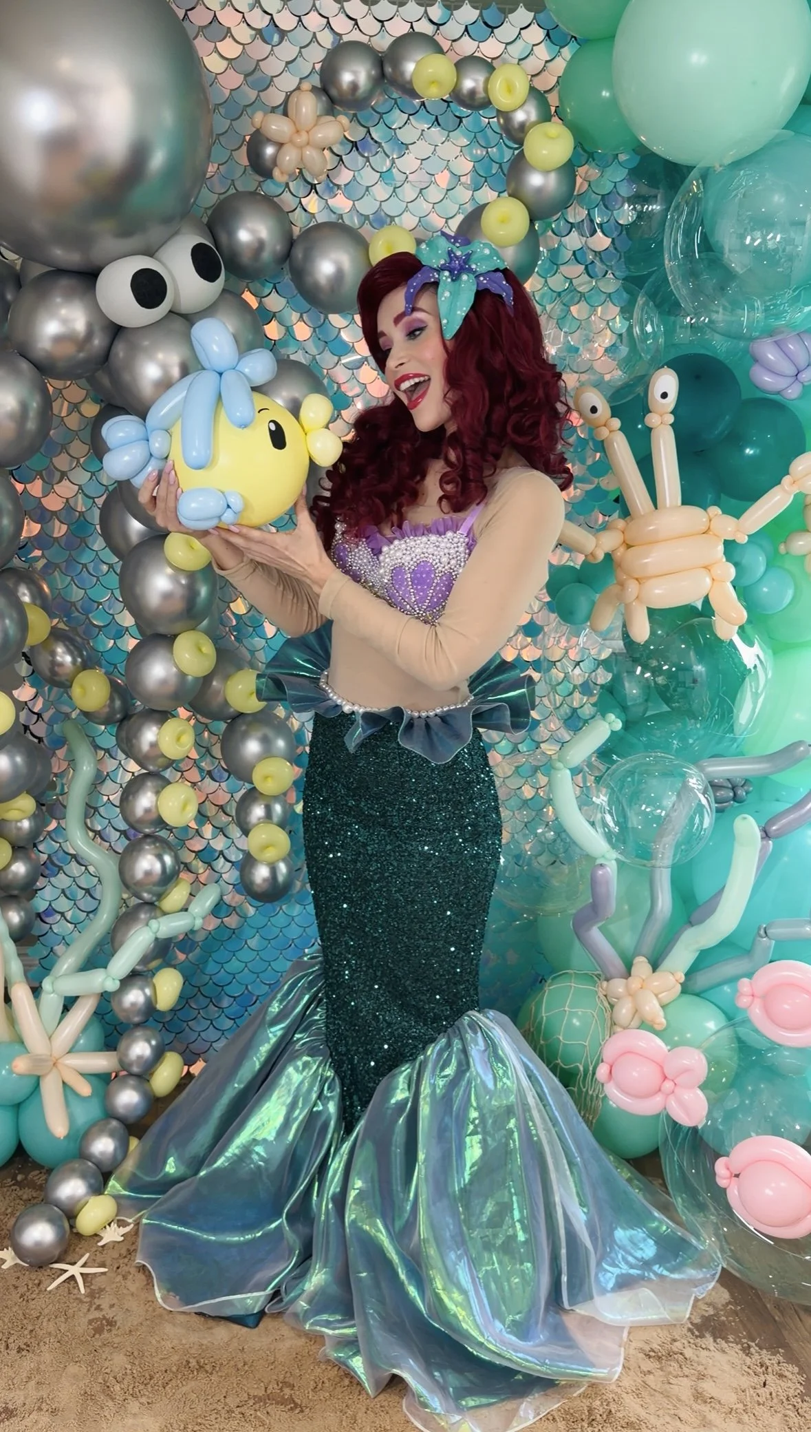 A woman in a mermaid costume with red hair and a colorful headband, holding a balloon crab and a balloon fish, surrounded by decorated balloons and an iridescent fabric skirt at an underwater-themed party setup.