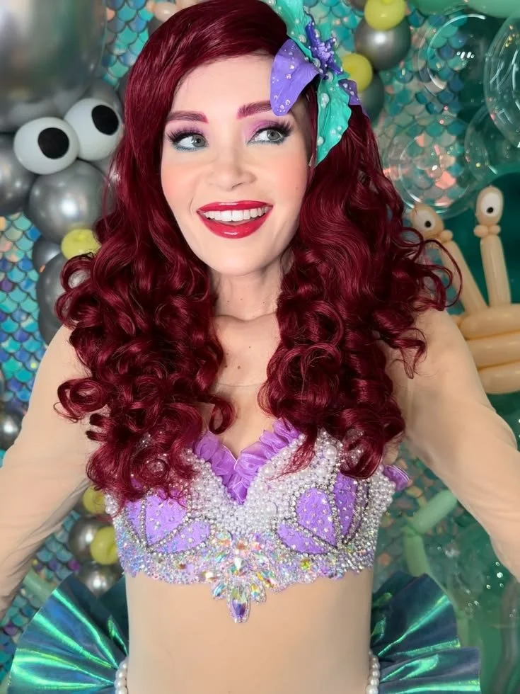 A woman with long, curly red hair and bright makeup, wearing a colorful purple and pearl embellished top with sheer beige sleeves, surrounded by balloons shaped like eyes and abstract decorations.