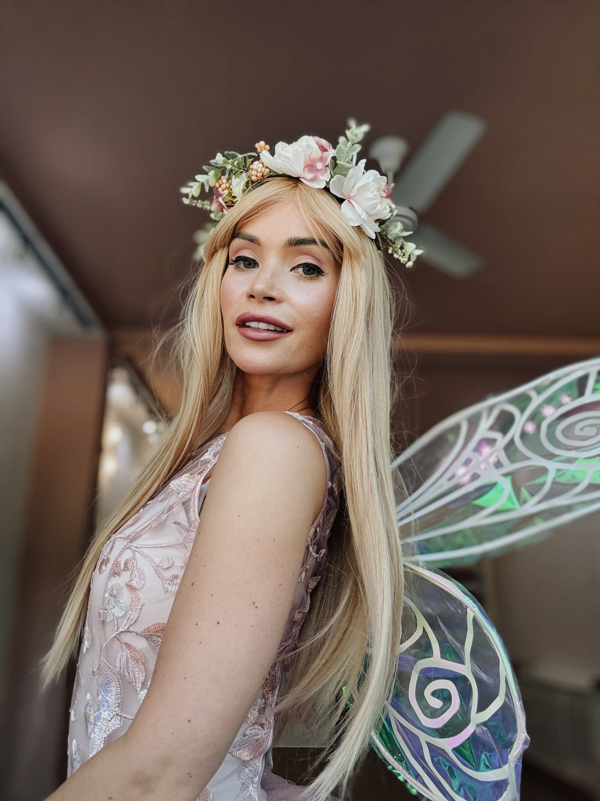 A woman with long blonde hair wearing a floral crown, pink embroidered dress, and fairy wings, smiling at the camera.