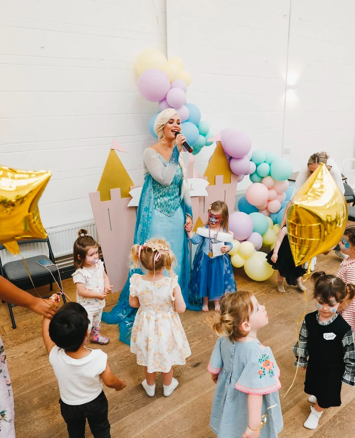 A children's birthday party with kids in colorful costumes, a woman dressed as Elsa from Frozen singing with a microphone, and pastel balloons in the background.