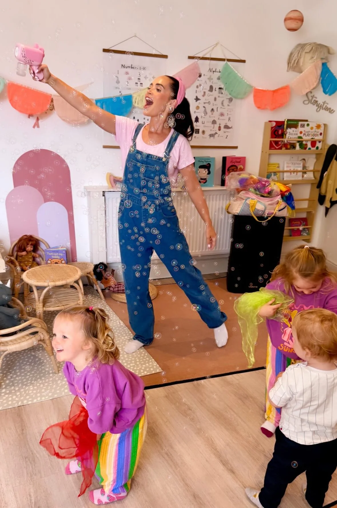 A woman in overalls and a pink headband is blowing bubbles in a children's party room decorated with colorful banners and posters. Several children are playing and interacting with the bubbles, with some wearing rainbow-colored skirts and others in c