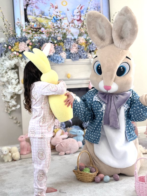 A child dressed in pajamas hugging a large yellow plush bunny. There is a person in a large bunny costume with big eyes and a polka-dot jacket, holding the child's hand. The background features pastel-colored Easter decorations, including stuffed bunnies and a floral display.