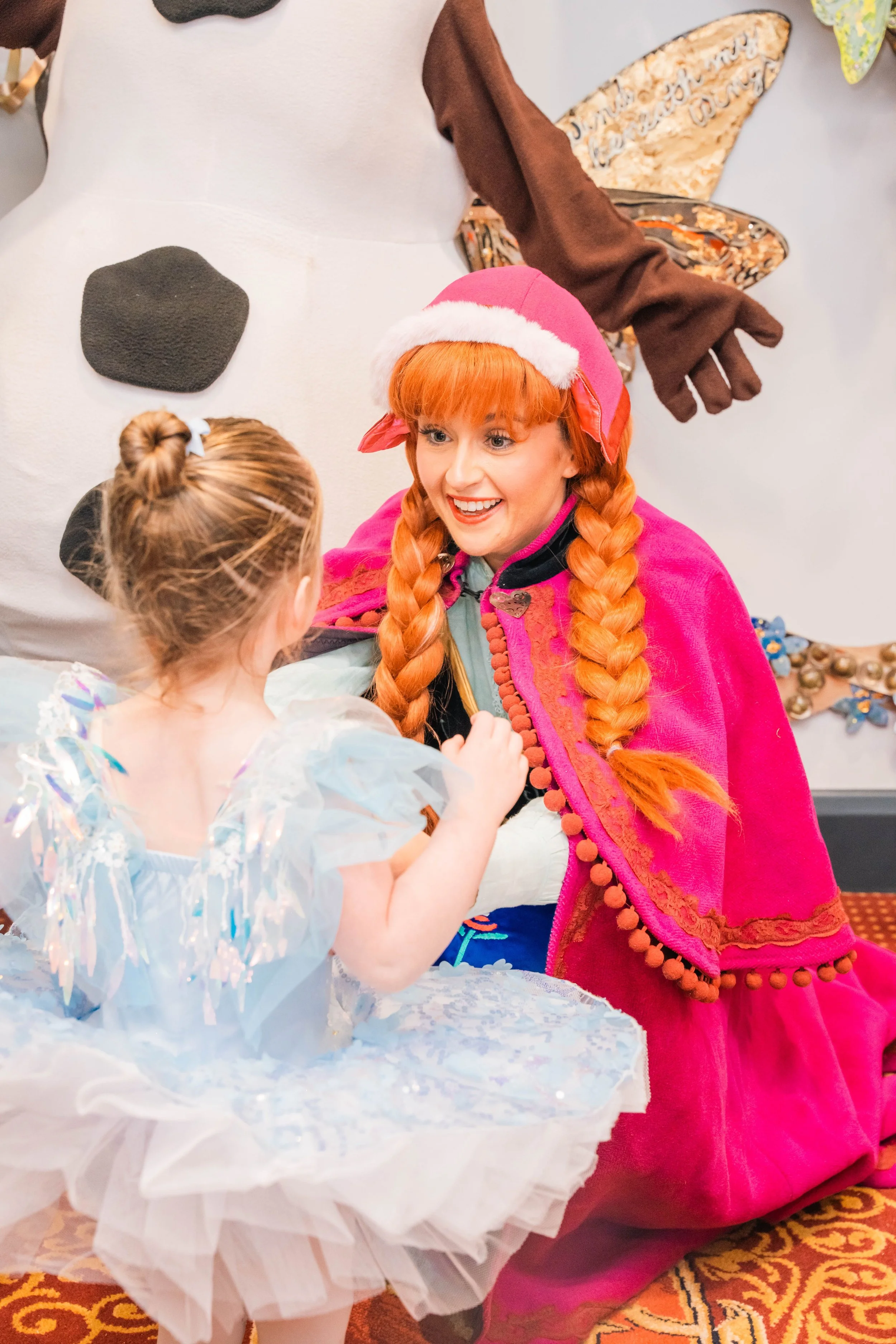 A woman dressed as a fairy godmother in a pink cape and Santa hat, smiling while talking to a young girl in a princess dress at a festive event.