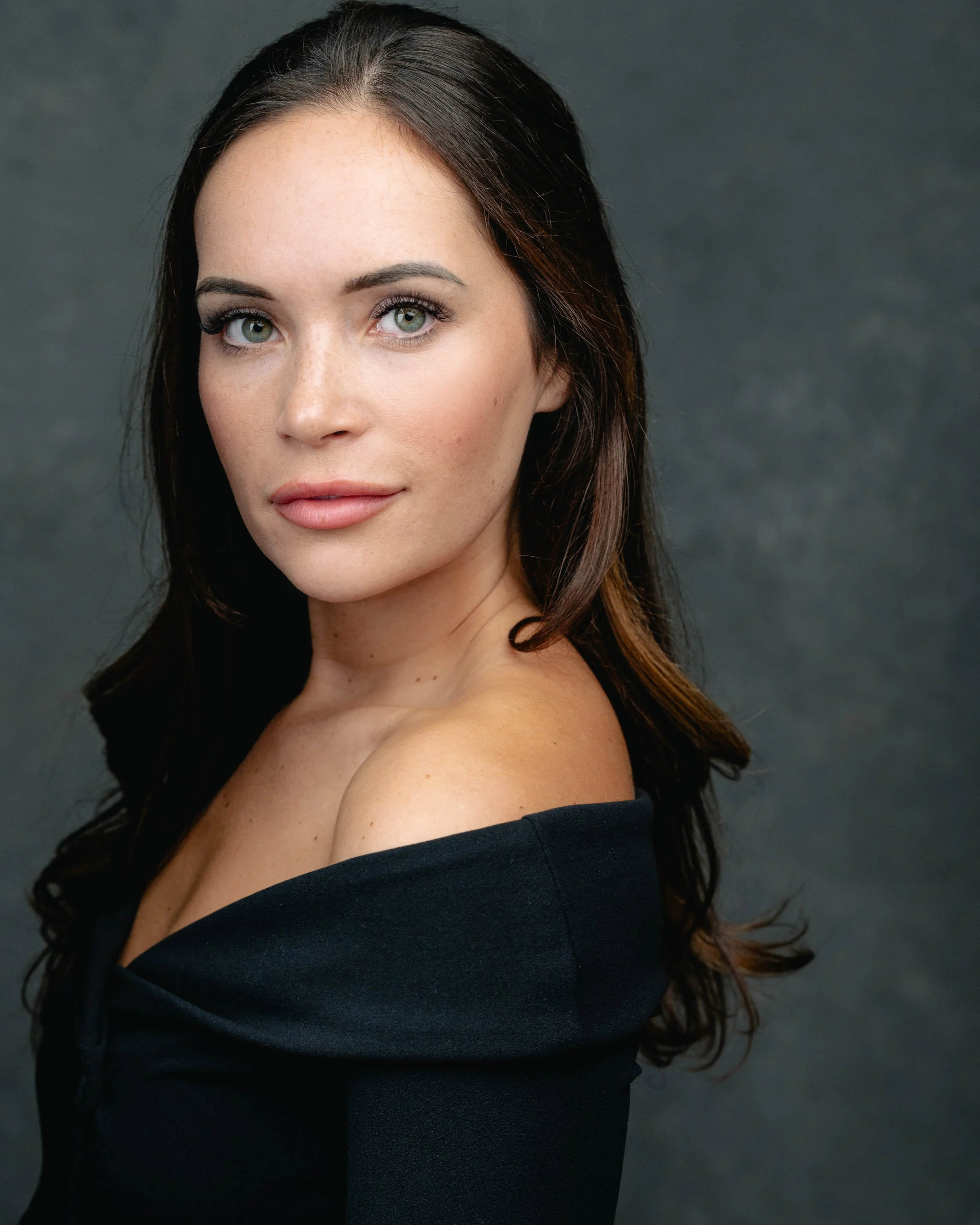 Close-up portrait of a woman with green eyes and long dark hair, wearing an off-the-shoulder black top, against a dark background.