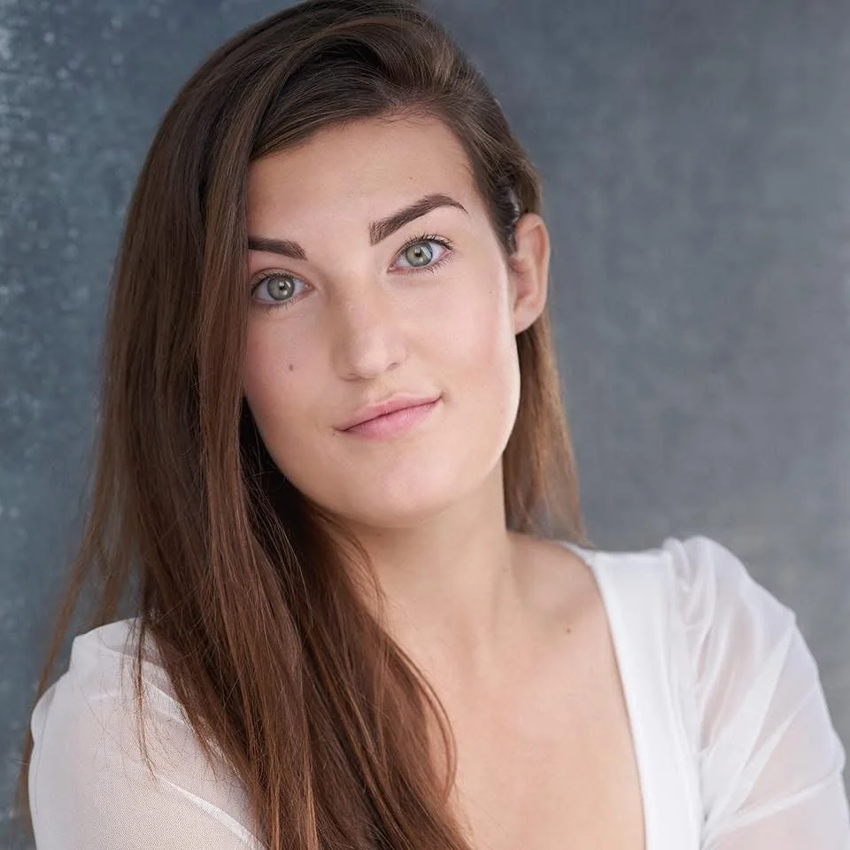 Close-up of a young woman with long brown hair, blue eyes, wearing a white top, and smiling softly against a gray background.