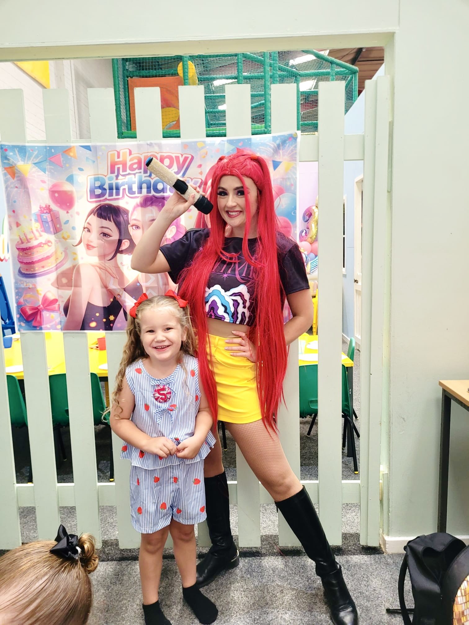 Young woman with long red hair wearing a black t-shirt, bright yellow shorts, and black thigh-high boots, standing next to a smiling young girl in a striped dress with red strawberry prints, at a birthday celebration with a 'Happy Birthday' banner an
