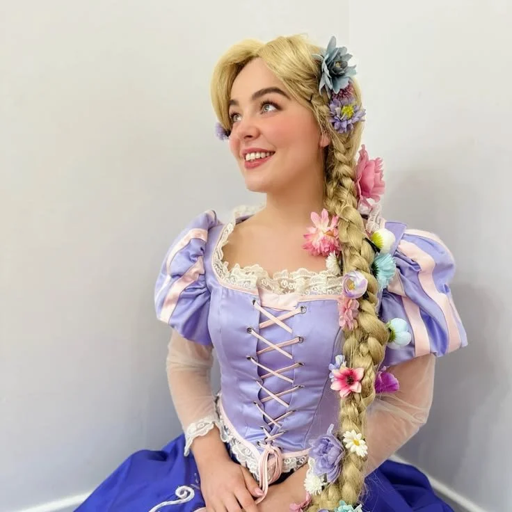 A woman dressed as Rapunzel with a pastel purple and pink costume, long braided blonde hair adorned with colorful flowers, standing against a plain white wall, smiling and looking off to the side.