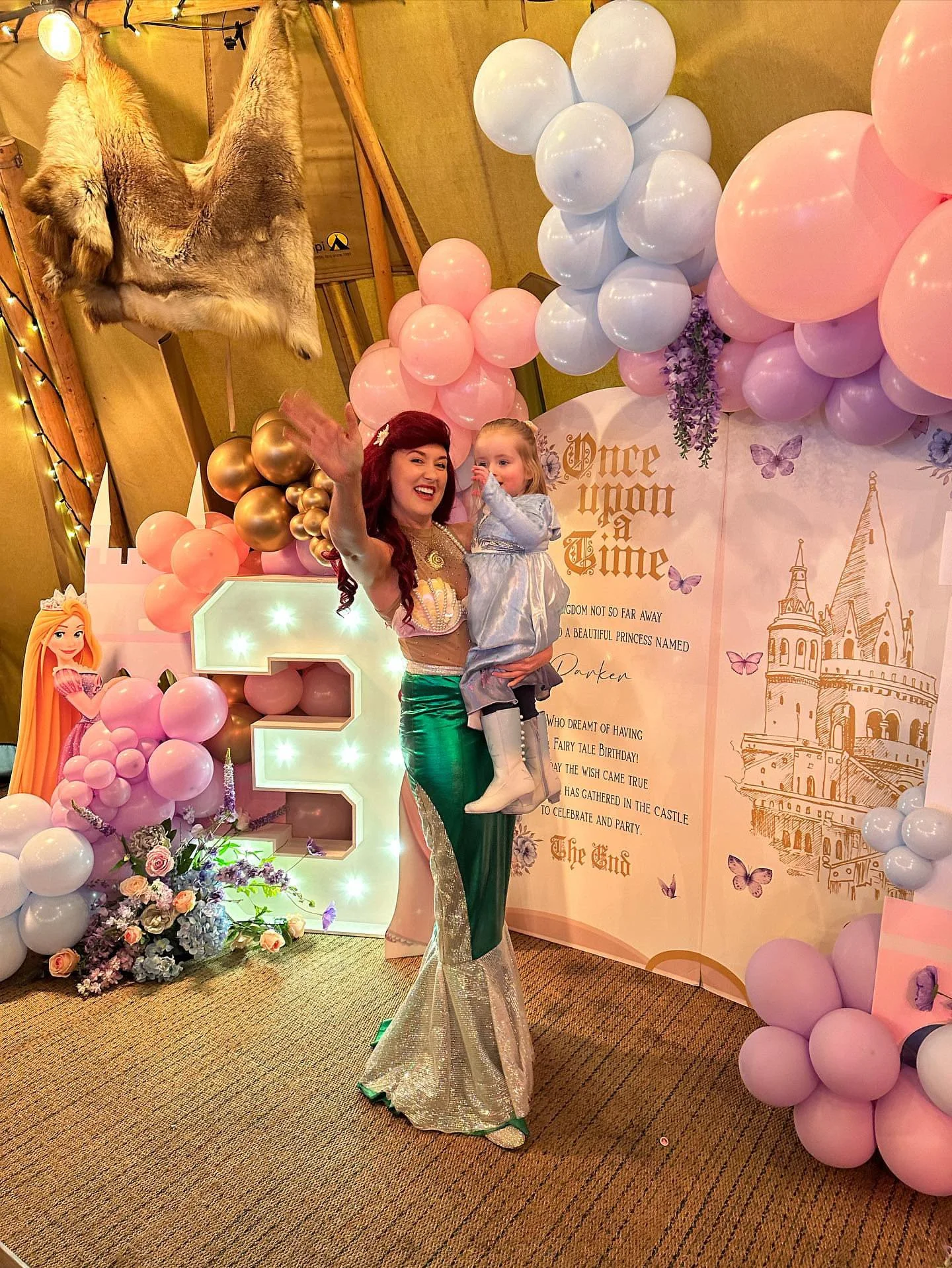 A woman dressed as a princess holding a young girl at a fairy tale birthday celebration with pink and white balloons, flowers, and castle-themed decorations.