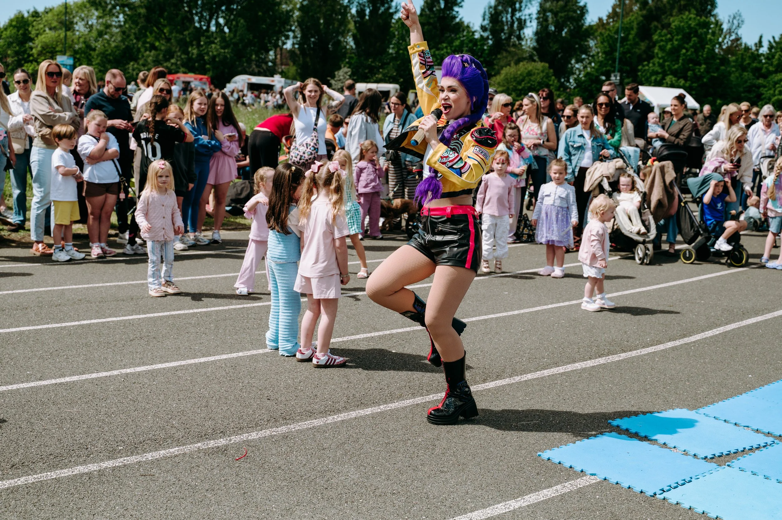 Street performer dressed in colorful costume performing at an outdoor event with a crowd of children and adults watching during daytime.
