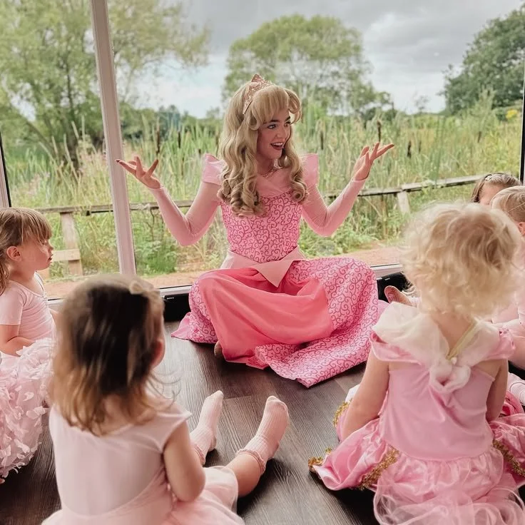 A woman dressed as a princess, sitting on the floor in pink, engaging with young children during a storytime or activity session in a bright room with large windows showing greenery outside.