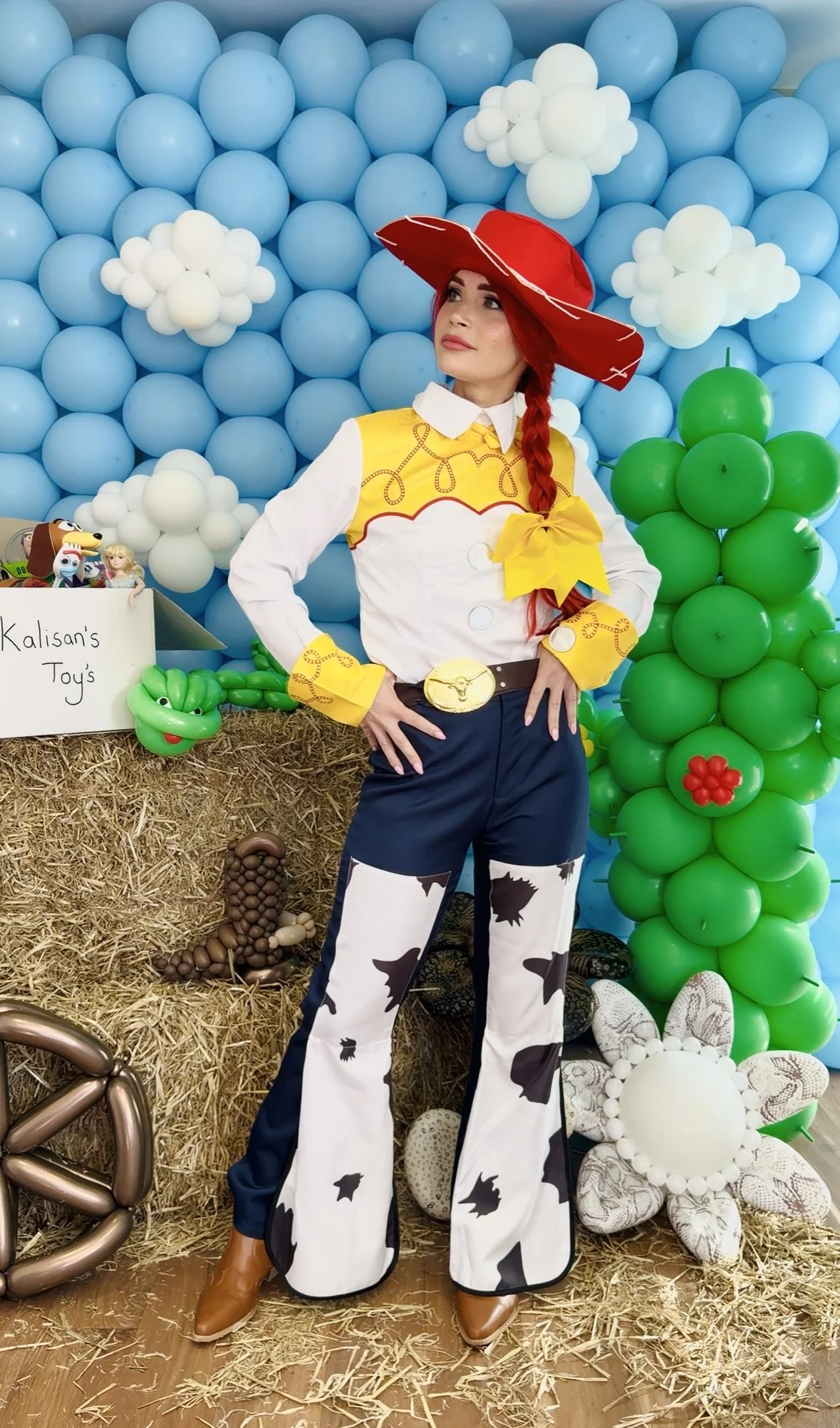 Woman in cowboy costume with red hat, yellow vest, and cow-patterned pants, standing in front of a backdrop made of blue and white balloons resembling clouds and sky, with a green balloon cluster and farm-themed decorations around.