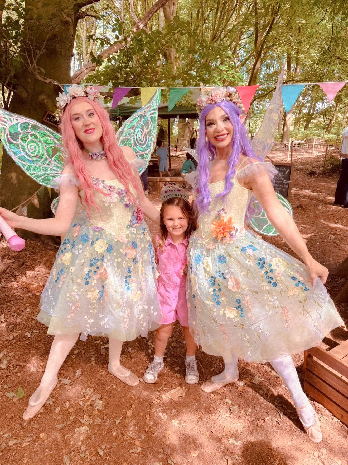 Two women dressed as fairies with pink and purple hair, fairy wings, and floral dresses, posing with a young girl in a pink jacket in an outdoor wooded setting decorated with colorful bunting, with other people in the background.