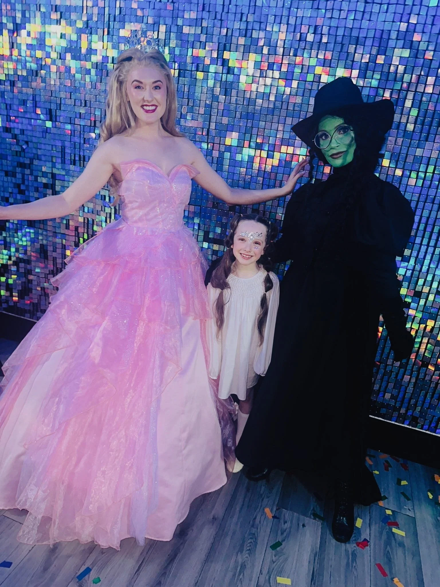 A woman dressed as a princess in a pink ball gown, a girl with face paint and a cream-colored dress, and another woman with green face paint, glasses, and a dark costume, standing in front of a colorful, mosaic-like tiled wall at a celebration or par