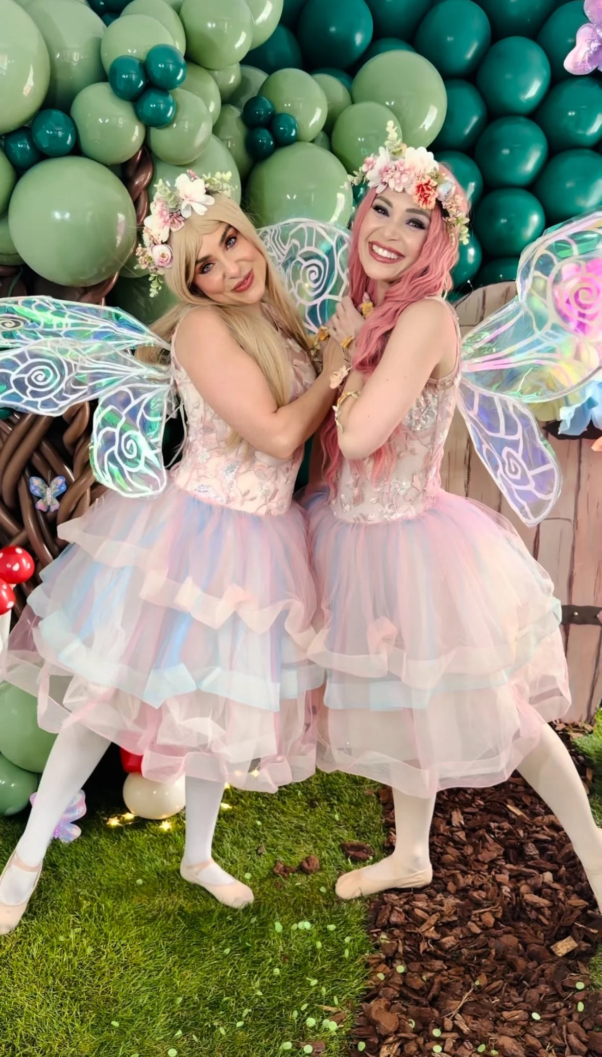 Two women dressed as fairies with pastel pink and blonde hair, wearing floral crowns, iridescent fairy wings, pink layered tutus, and white tights, standing in front of a background of green and teal balloons and fairy garden decorations.