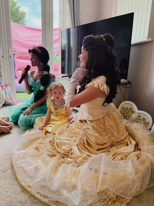 Two women dressed as princesses and a young girl sitting on a quilted blanket on the floor during a princess-themed party, with a television and window in the background.