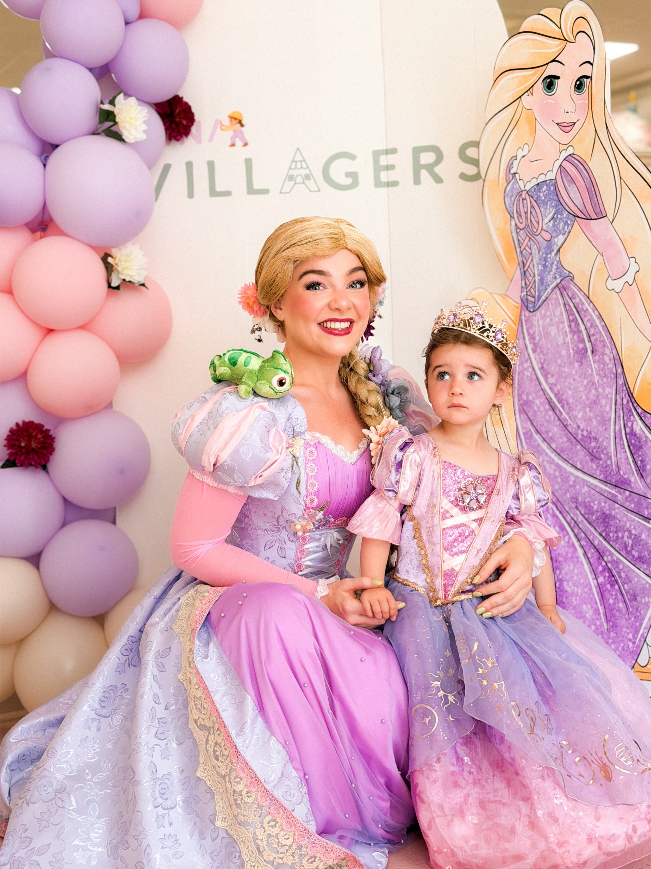 A woman dressed as Rapunzel from Disney's Tangled, with a young girl in a princess costume, both seated in front of a Rapunzel-themed backdrop decorated with pink and purple balloons and flowers.