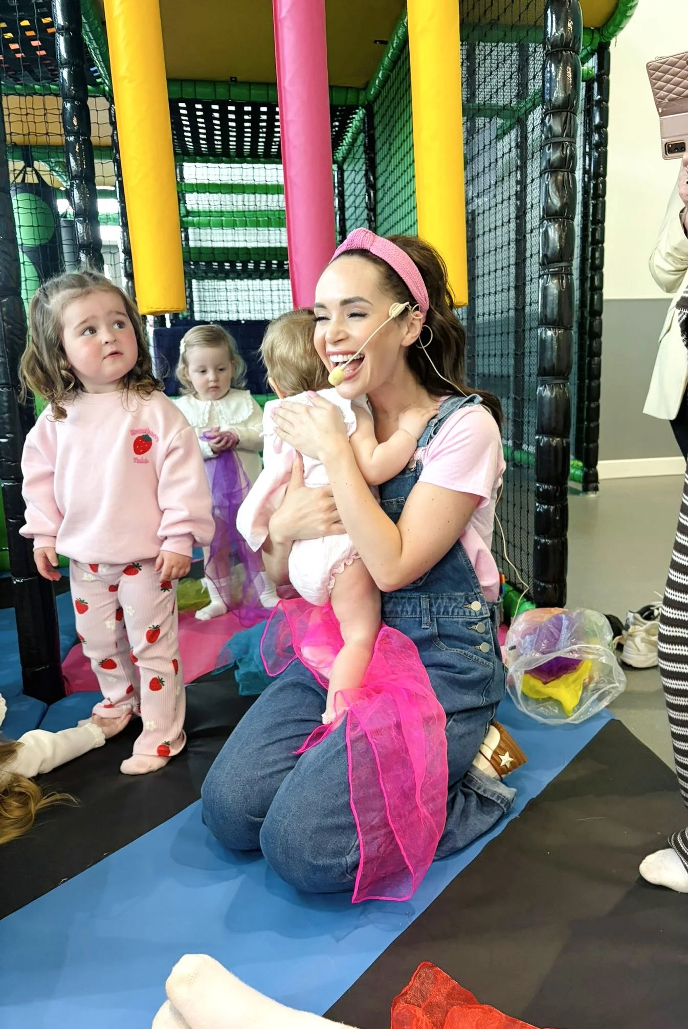 Smiling woman with a pink headband holding a young girl in a pink tutu at an indoor play area, with several children around, including a girl in strawberry pajamas and a girl in a princess dress, surrounded by colorful padded play structures.