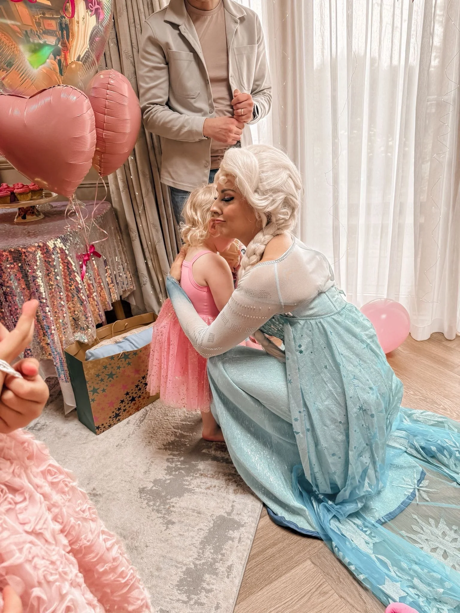A woman dressed as Elsa from Frozen hugging a young girl in a pink dress at a party with pink balloons and gifts.
