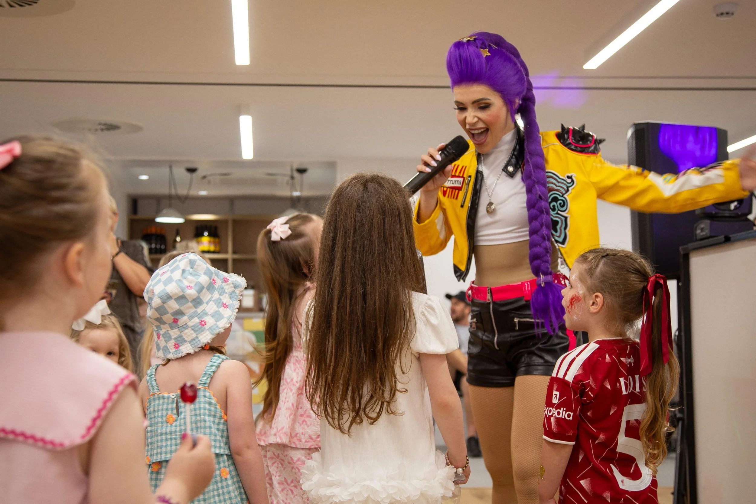 A performer with purple hair styled in a long braid, wearing a yellow jacket and black shorts, is singing into a microphone while engaging with children in a room with a modern ceiling and ambient lighting.