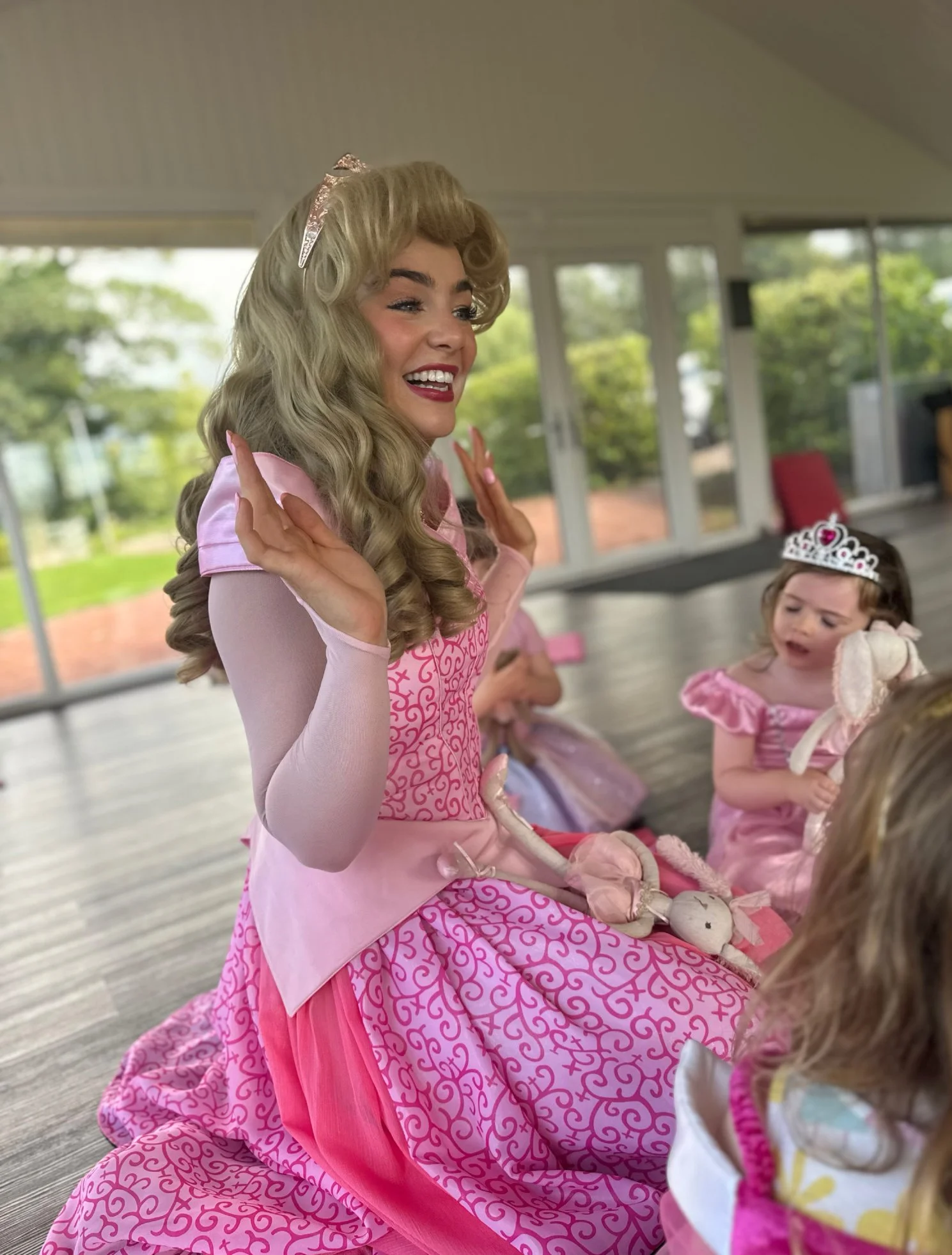 A young woman dressed as a princess with a long pink dress with heart patterns, a shiny blonde wig, and a tiara, smiling and gesturing with her hands while sitting with children dressed as princesses in a bright indoor space with large windows and gr