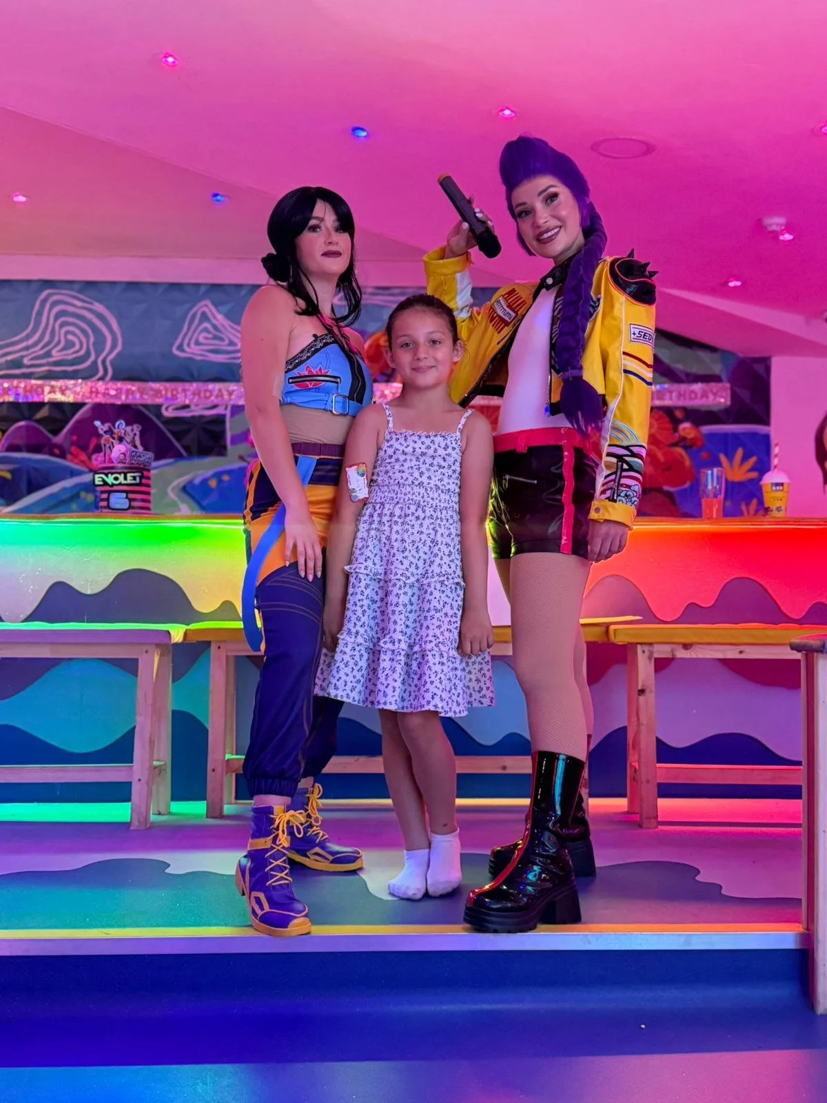 Two women in colorful outfits and a young girl standing in a neon-lit arcade or entertainment venue. One woman is holding a toy gun, and they are all smiling.