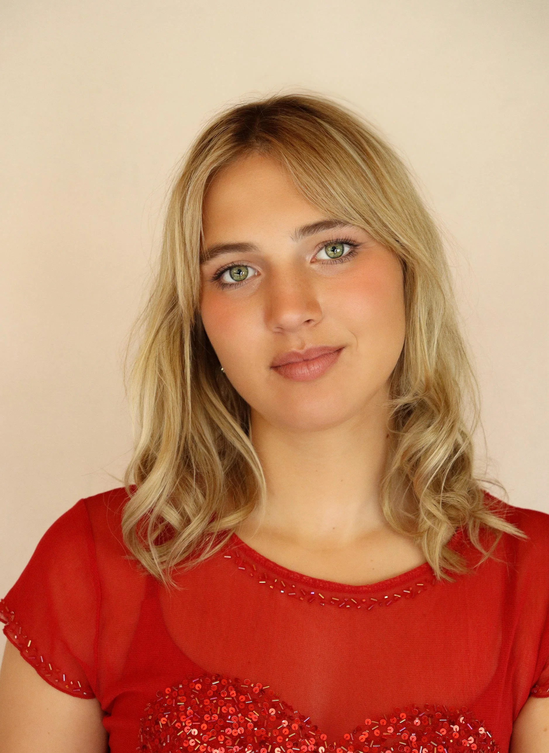 Portrait of a young woman with blonde, wavy hair and green eyes, wearing a red top with sequin embellishments around the neckline and sleeves, standing against a plain beige background.