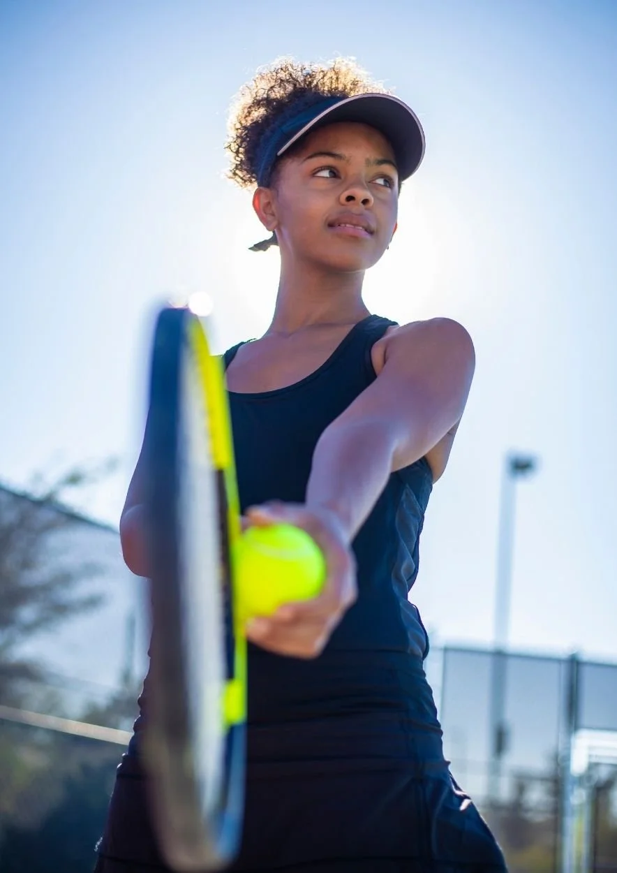 Jeune femme noire jouant au tennis sur un court extérieur, portant une casquette, t-shirt noir et tenant une raquette de tennis, avec le soleil en arrière-plan.