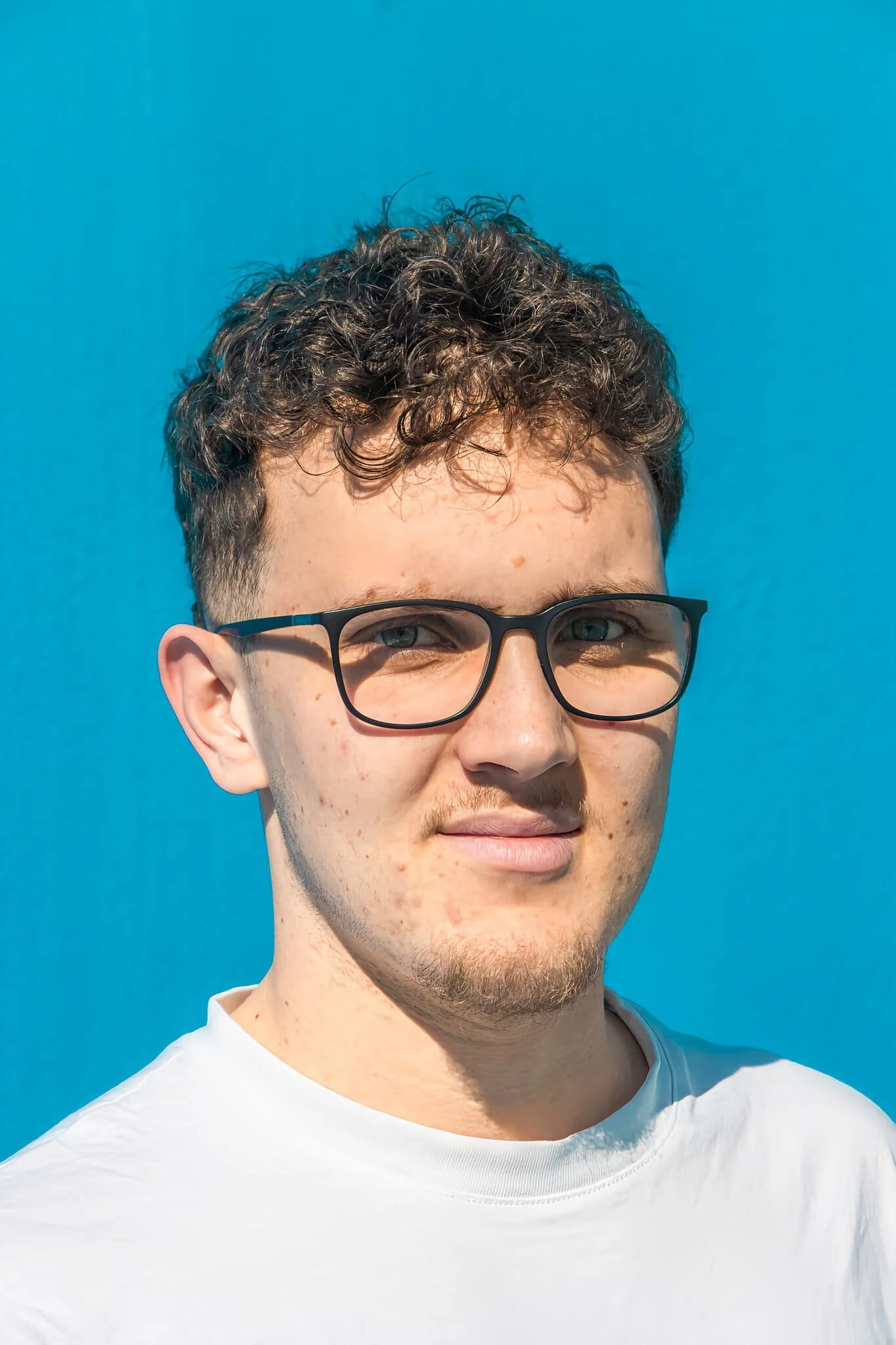 Jeune homme avec cheveux bouclés et lunettes, souriant, portant un t-shirt blanc, contre un fond bleu clair.