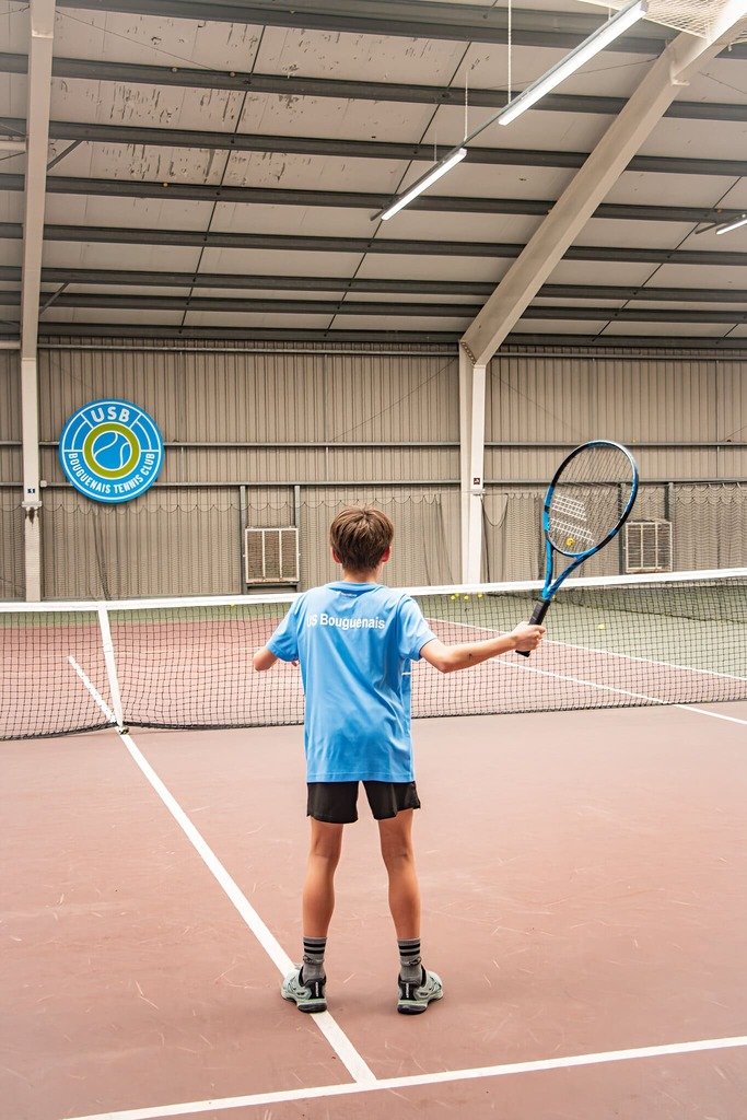 Un jeune garçon en t-shirt bleu et short joue au tennis à l'intérieur d'un court couvert.