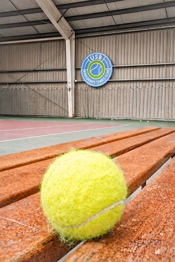 Une balle de tennis jaune posée sur un banc en bois dans une salle de tennis en intérieur, avec un mur en arrière-plan portant un panneau bleu indiquant "USC Bouquena Tennis Club".