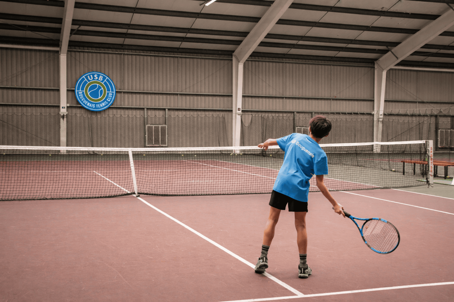 Un jeune garçon jouant au tennis dans une salle intérieure, portant un t-shirt bleu avec le nom du club "US Bougnenais" et tenant une raquette, avec un filet de tennis devant lui.