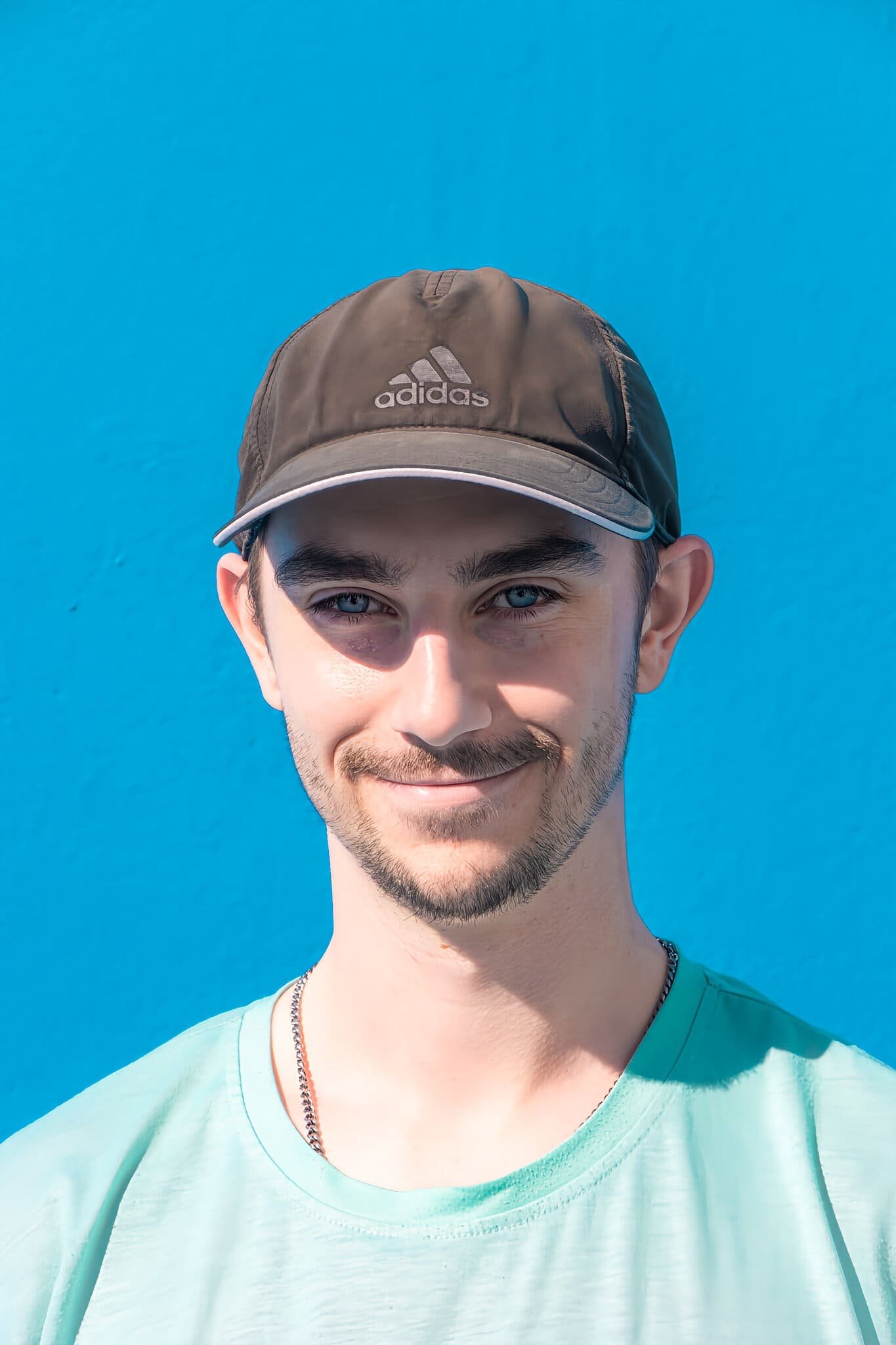 Jeune homme souriant portant une casquette Adidas et un t-shirt vert clair, avec un fond de ciel bleu.