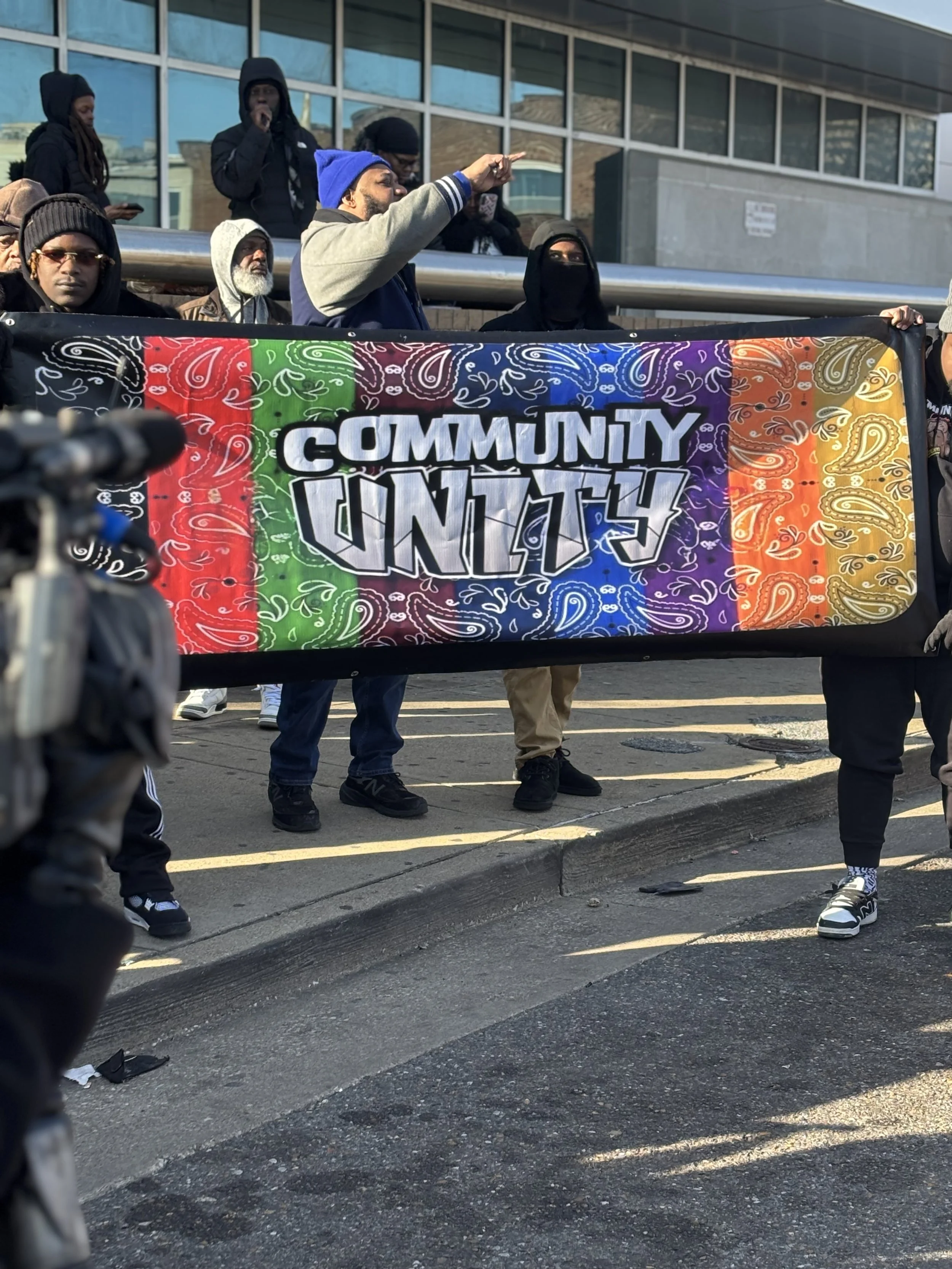 A diverse group of people holding a banner that says 'Community Unity' with a rainbow-colored background and a paisley pattern, participating in a street protest or march.