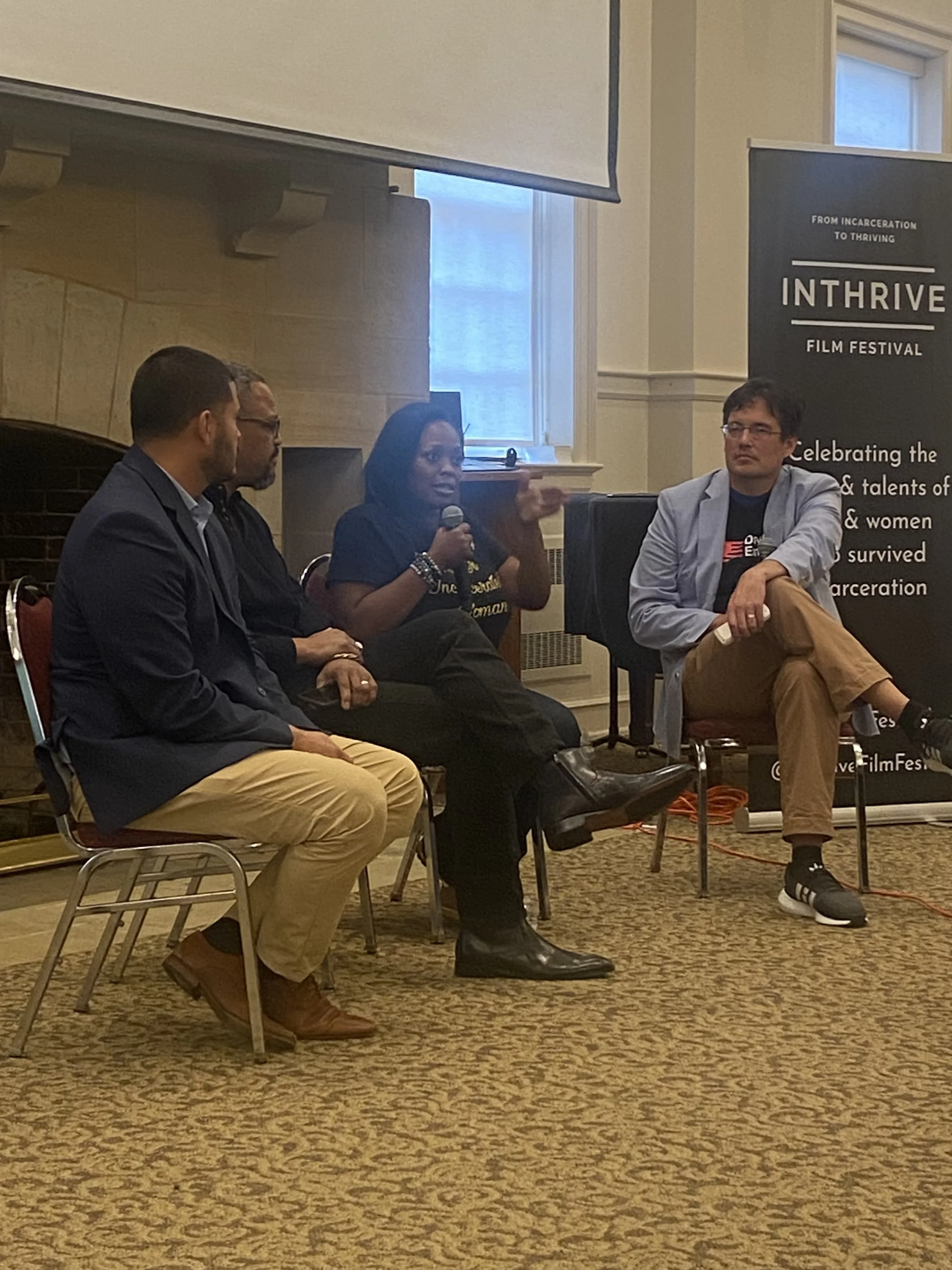Four individuals sitting in chairs during a panel discussion at the Inthrive Film Festival, with one woman speaking into a microphone, the festival banner in the background, and a window letting in natural light.