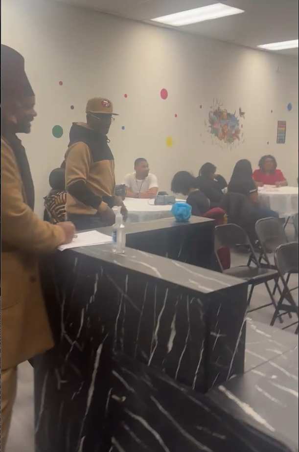 People gathered in a room with tables and chairs, some seated and some standing, with colorful wall decorations including polka dots and artwork.