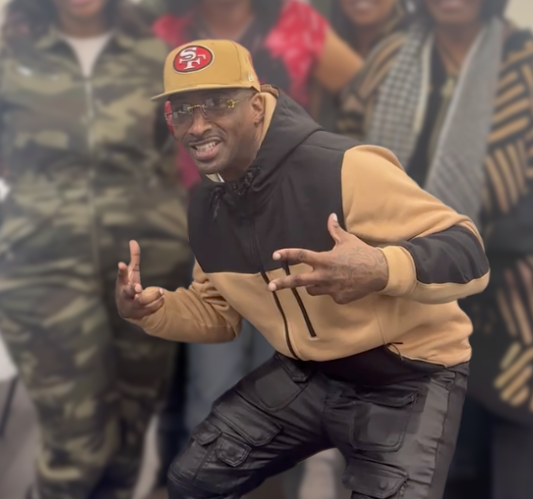A man wearing a tan and black hoodie, black cargo pants, sunglasses, and a tan cap with a sports logo, making peace signs and smiling, with a group of people in the background.