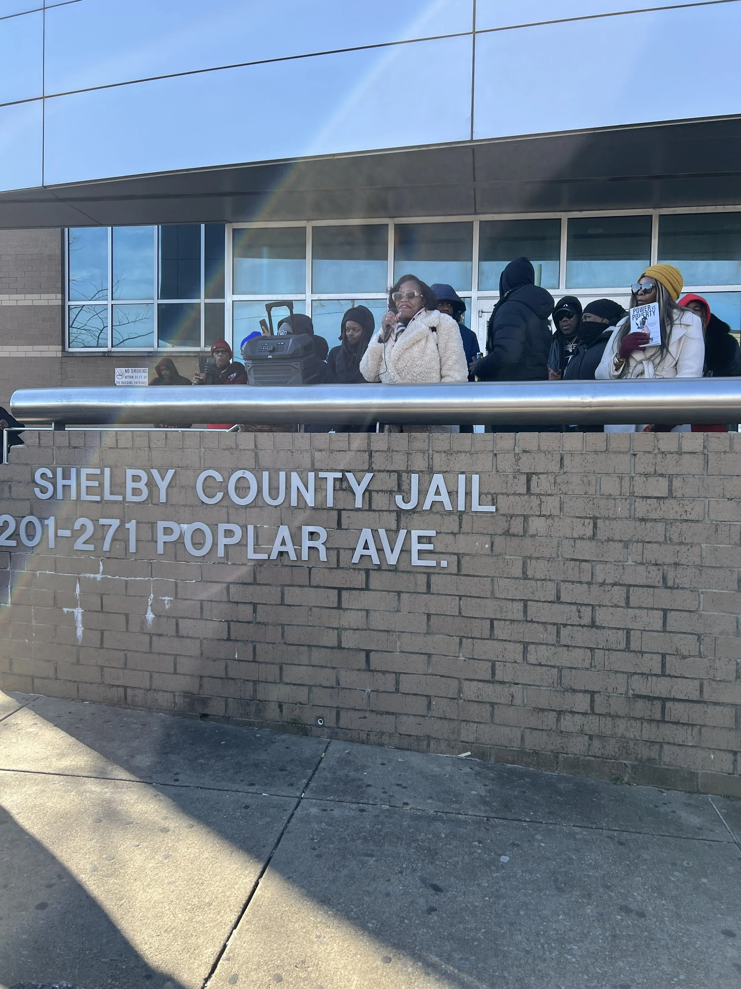 People protesting outside Shelby County Jail, some holding signs, with a woman speaking into a microphone, in front of a brick wall and a building with glass windows.