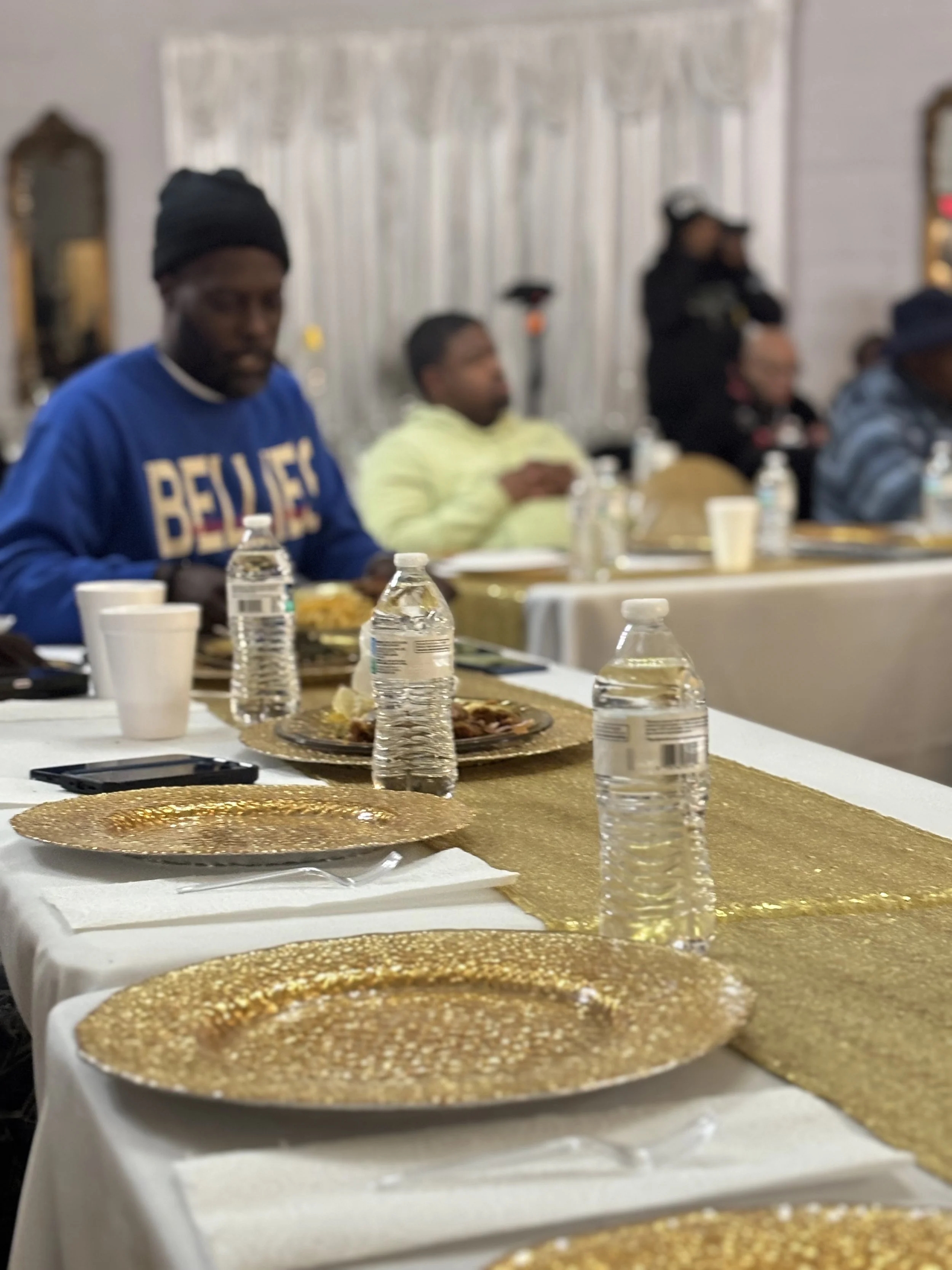 People sitting at a table with gold tablecloths, water bottles, and plates of food in a banquet hall.