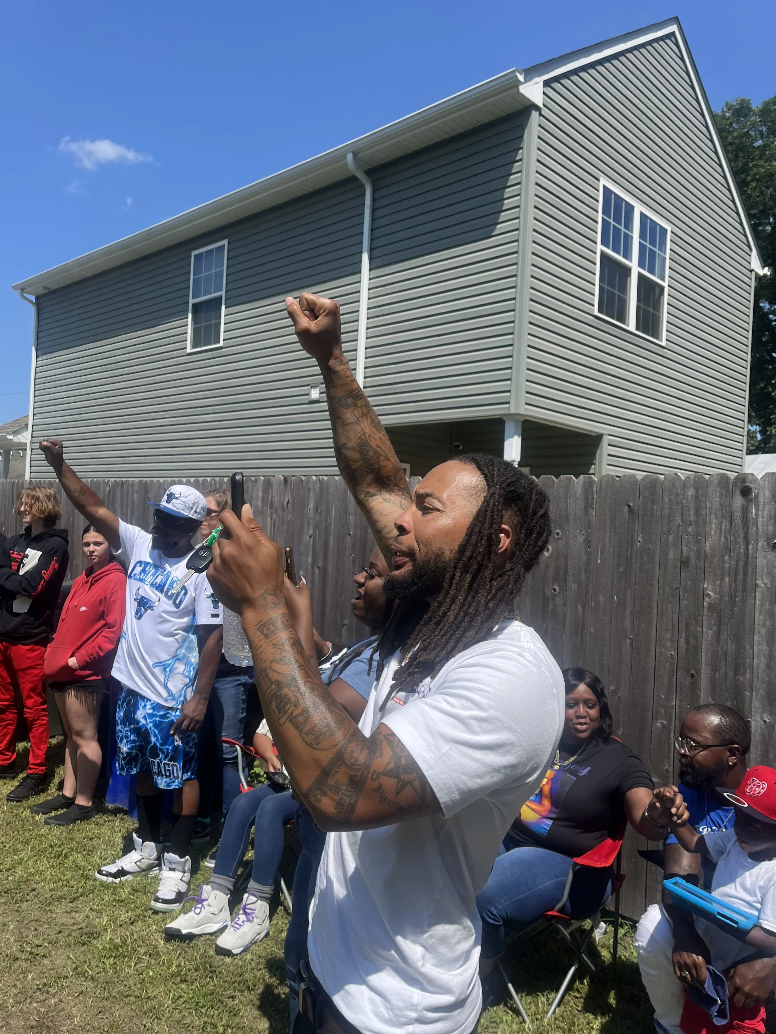 A group of people standing outdoors behind a wooden fence, raising their fists, with a gray house and blue sky in the background.