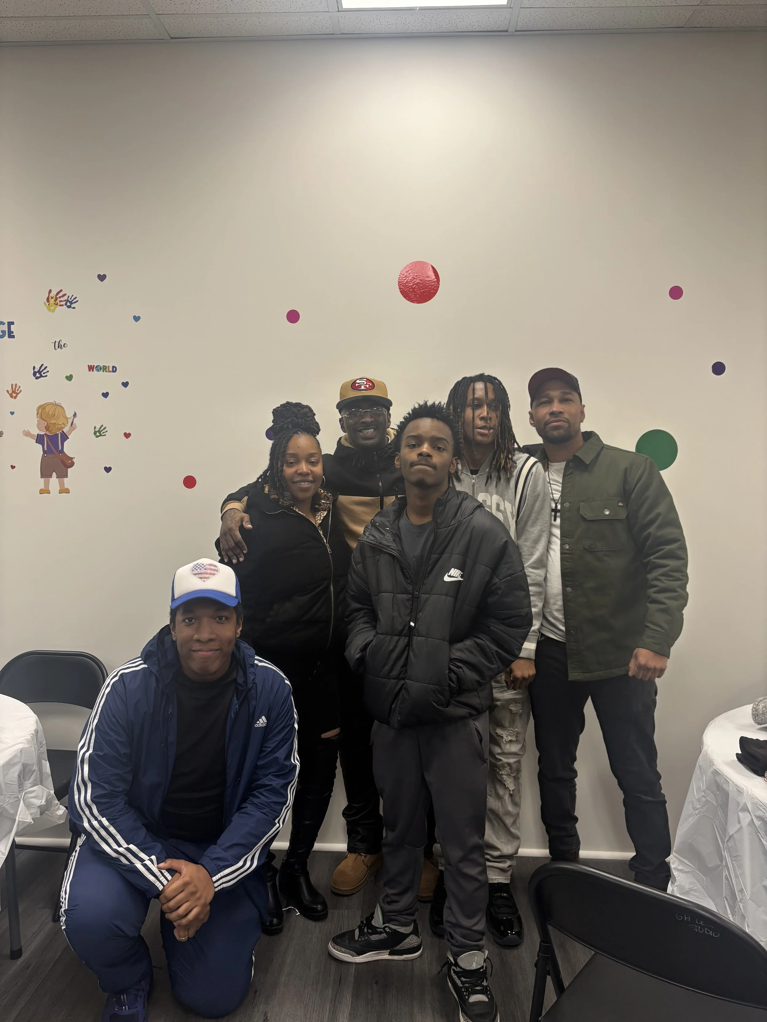 Group of five people posing for a photo in a room with decorated wall. One person is kneeling in front, four are standing behind.
