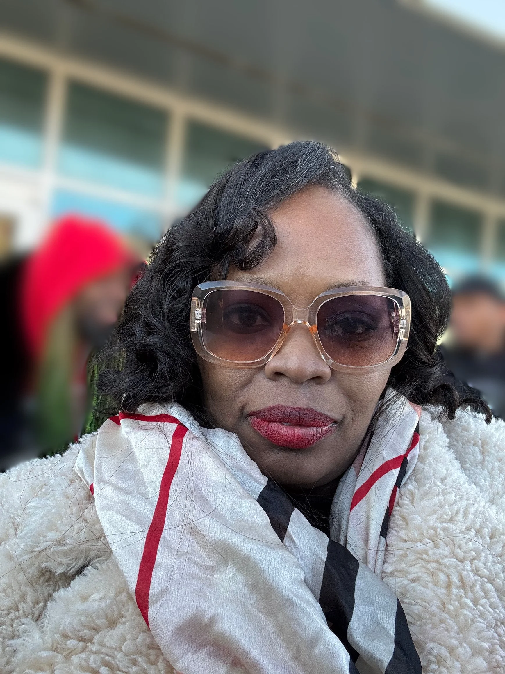 Close-up of a woman wearing large sunglasses and a white jacket with red accents, sitting on a fluffy surface outdoors with a blurred building in the background.
